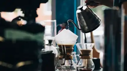 A gooseneck kettle pouring over a glass brewer on the roastery counter.